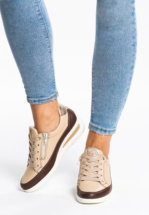 Person wearing beige and brown sneakers with side zippers and light blue skinny jeans against a white background.