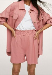 Femme portant un short rose ample et une veste à boutons assortie sur un T-shirt blanc, les mains sur les hanches.