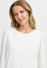 Vit texturerad långärmad topp med ett vågigt randigt mönster. Halsringningen är rund och har en mjuk finish. Subtil lyster synlig.