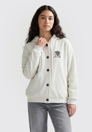 Kvinde med langt mørkt hår iført en hvid hættetrøje med knapper og et våbenskjoldspatch samt grå jeans, stående foran en ensfarvet lys baggrund.