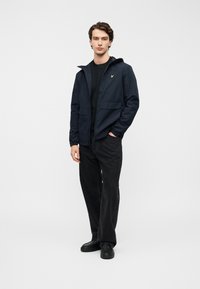 Ναυτικό μπλε μπουφάν με κουκούλα, μπροστινές τσέπες, κλείσιμο με φερμουάρ και ελαστικά μανίκια, φορεμένο πάνω από μαύρο πουκάμισο και συνδυασμένο με μαύρο παντελόνι και παπούτσια.