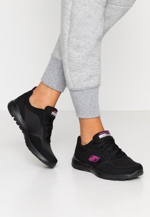 Des chaussures de sport noires en matériaux synthétiques et en maille, avec un logo rose, des lacets et une semelle texturée. Associées à un pantalon de survêtement gris clair.