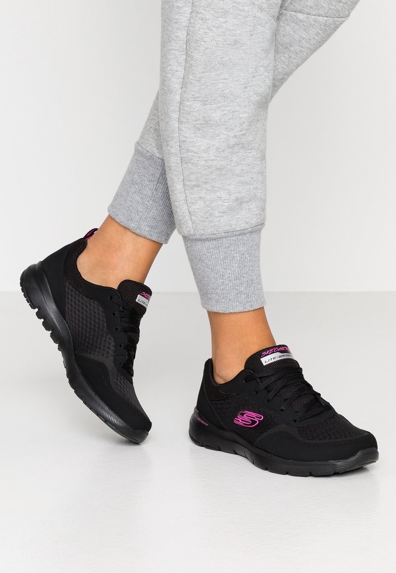 Scarpe da ginnastica nere realizzate in mesh e materiali sintetici, con un logo rosa, lacci e una suola testurizzata. Abbinate a pantaloni della tuta grigi chiari.