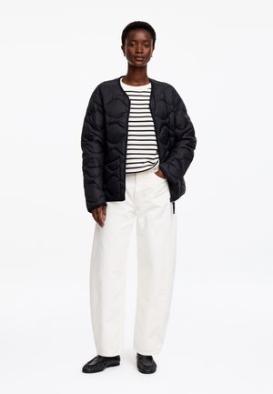 Femme portant une veste matelassée noire, une chemise rayée noir et blanc, un pantalon blanc à jambes larges et des chaussures noires, debout sur fond blanc.