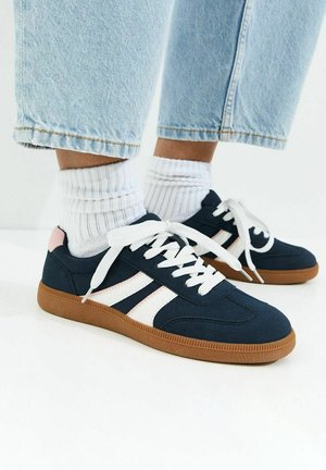 Stopy w granatowych trampkach z białymi sznurowadłami i paskami, białe skarpetki oraz jasnoniebieskie podwinięte dżinsy na białym tle.