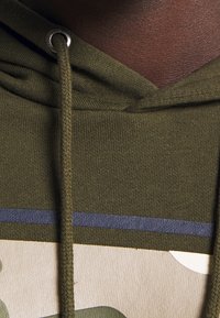 Sudadera color verde oliva con un tejido texturizado, con una franja azul en la parte frontal y un cordón con ojales de metal.