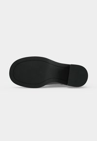 Semelle de chaussure noire avec une surface lisse légèrement texturée et un talon distinct, vue directement de dessous sur un fond blanc.