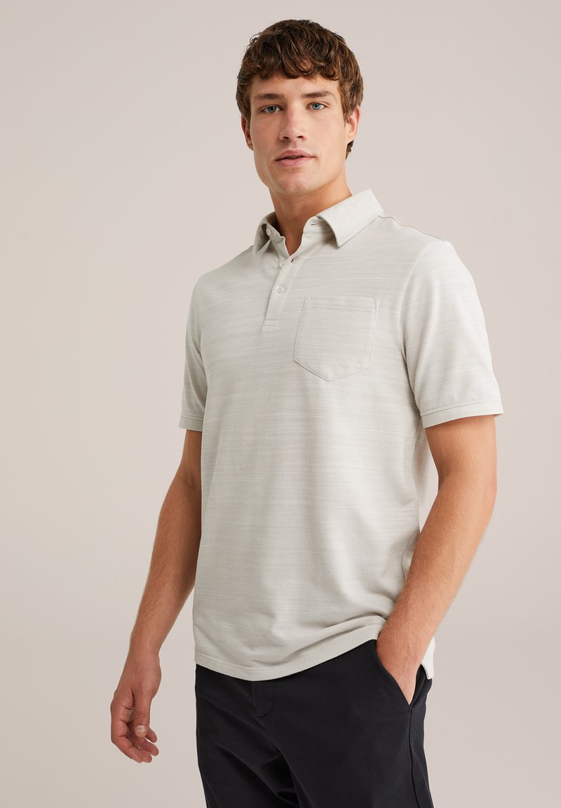 Joven con cabello corto marrón, vestido con una camiseta polo de manga corta color beige claro y pantalones negros, de pie con una mano en el bolsillo.