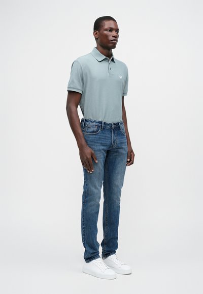 Ανοιχτό πράσινο πόλο μπλουζάκι με διπλωμένο γιακά, συνδυασμένο με σκούρες μπλε τζιν παντελόνες ίσιας γραμμής. Άσπρα sneakers ολοκληρώνουν την εμφάνιση. Μινιμαλιστικός σχεδιασμός.