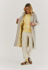 Dlhý sivý kabát cez žltú mikinu s doškrabaným textom, krémové nohavice a tenisky s leopardím vzorom a bielymi akcentmi.