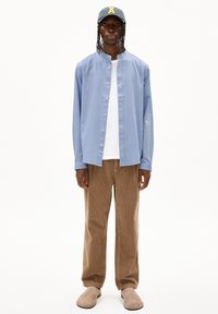 Helltblaues Hemd mit Knöpfen, darunter ein weißes T-Shirt, braune Cordhosen und beiges Slipper, ergänzt durch eine marineblaue Mütze mit gelben Akzenten.