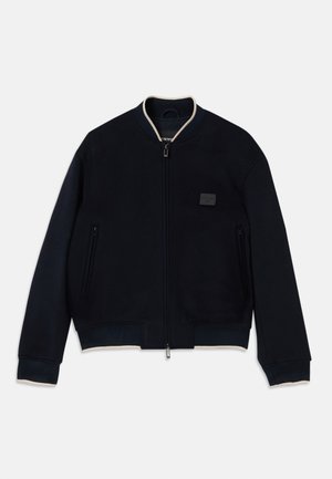 Navy bomber jacket made of soft fleece with a zip closure. Features a contrasting beige collar, side pockets, and a textured logo patch.