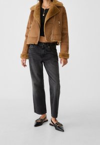 Giacca in shearling color tan con un design cropped e chiusura zip, abbinata a jeans neri a vita alta con gamba dritta e scarpe slingback nere basse.