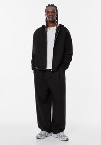 Schwarze Kapuzen-Zipfeljacke über einem weißen Rippenshirt, kombiniert mit schwarzen weiten Hosen und hellgrauen Turnschuhen. Glattes Material, lockere Passform.