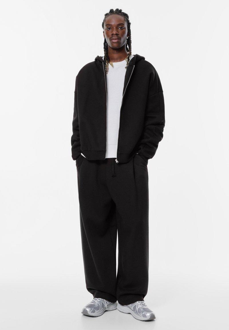 Schwarze Kapuzen-Zipfeljacke über einem weißen Rippenshirt, kombiniert mit schwarzen weiten Hosen und hellgrauen Turnschuhen. Glattes Material, lockere Passform.
