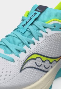 Λευκό και τυρκουάζ αθλητικό παπούτσι με τυρκουάζ κορδόνια, επενδυμένο γλώσσα με ετικέτα "Saucony" και λογότυπο της μάρκας με κίτρινες λεπτομέρειες στο πλάι.