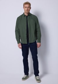 Camisa de cuadros verdes con un bolsillo en el pecho, usada sobre una camiseta negra, combinada con jeans oscuros y zapatillas verdes.