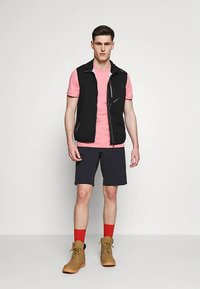 Nuori mies seisoo päällä musta vetskari, vaaleanpunainen t-paita, mustat shortsit, punaiset sukat ja vaaleanruskeat nauhalla kiinnitettävät saappaat pelkän taustan edessä.