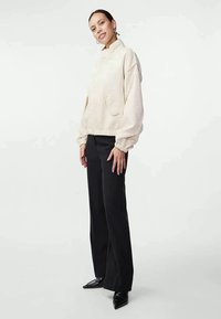 Pullover cortado de cor creme com colarinho alto, grandes bolsos e punhos elásticos, combinado com calças pretas de corte ajustado e sapatos de bico.
