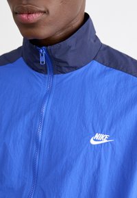 Zila jaku ar rāvējslēdzēju un augstu apkakli; ar tumši ziliem akcentiem un baltu Nike logo uz krūtīm. Strukturēta auduma materiāls ar mūsdienīgu piegriezumu.
