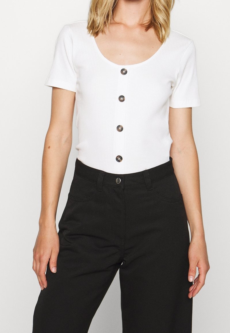 Haut blanc côtelé à manches courtes avec un col rond et cinq boutons foncés sur le devant, associé à un pantalon noir taille haute.