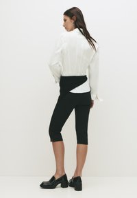 Witte blouse met lange mouwen en verticale strepen, zwarte cropped leggings met zijsplitten, en glanzende zwarte hakloafers.