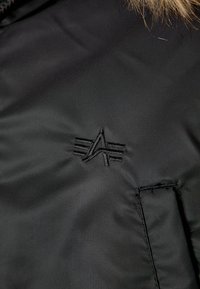 Sort isoleret jakke med en blød tekstur, broderet logo med skarpe linjer og en lomme detaljeret på stoffet.