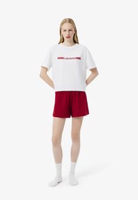T-shirt bianco a maniche corte con logo rosso 'LACOSTE', abbinato a pantaloni corti rossi. Modello che indossa calzini bianchi alla caviglia, braccia rilassate ai lati.
