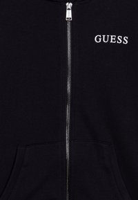 Černá mikina s kapucí na zip a předními kapsami, s stříbrným zipem a bílým vyšitým logem "GUESS" na hrudi. Měkká textilie.