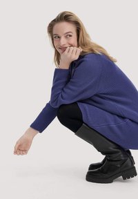 Maglione viola a maniche lunghe e a costine abbinato a leggings neri e stivali neri fino al ginocchio. La persona è accovacciata e sorride leggermente.