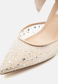 Escarpin beige à bout pointu, talon haut avec bride à la cheville et tissu transparent orné de petits et moyens strass argentés.