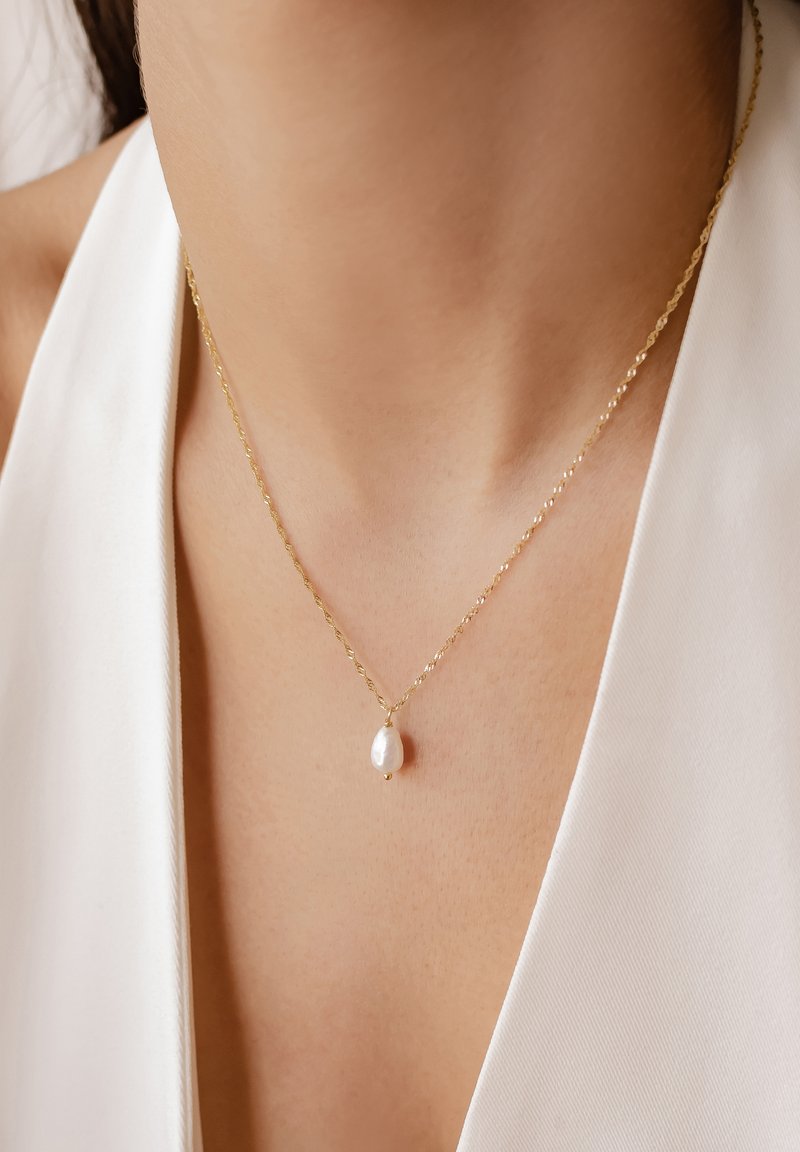 Złoty naszyjnik z skręconym łańcuchem i łezkowatym białym perłowym wisiorkiem, spoczywający na gładkiej skórze na tle białej tkaniny.