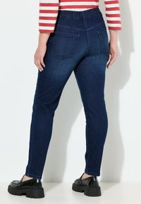 Žena v tmavě modrých úzkých džínech s elastickým pasem, černých silných mokasínech a dlouhém tričku s červenými a bílými pruhy, stojící.