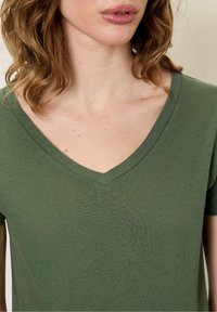 T-shirt verde oliva a V realizzato in un tessuto morbido. Presenta maniche corte e una texture liscia con delicate cuciture lungo il colletto.