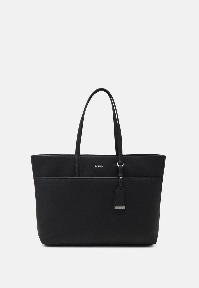 Sac cabas noir Calvin Klein avec deux poignées et une étiquette suspendue, présenté sur un fond gris clair uni.