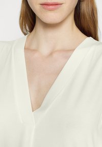 Blouse couleur crème avec un décolleté en V profond, texture douce et coupe ample, avec des coutures minimales et sans embellissements visibles.