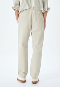 Helle beige Baumwollhose mit lockerer Passform, elastischem Bund und Taschendetails auf der Rückseite. Kombiniert mit weißen Schuhen und einem gestreiften Hemd.