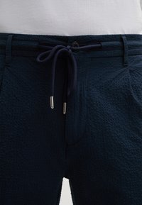 Pantaloni blu navy con texture e vita con cordoncino, dotati di estremità in metallo argentato, dettagli delle tasche discreti e un tessuto a fantasia traspirante.