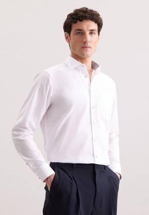 White long-sleeve shirt with a classic collar, button-up front, and single chest pocket; paired with dark trousers showcasing a tailored fit.