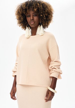 Lichtroze gebreide trui met textuur en diagonale patronen, oversized mouwen. Accessoires zijn grote witte kwastoorbellen en een crème armband.