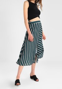 Teal kjol med vita vertikala ränder, medellång, lätt tyg, med en omslagsdesign och en sidoklyvning. Svart crop top och skor.