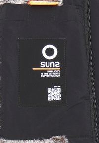 Etichetta in tessuto nera con logo circolare bianco e testo; codice QR sottostante. Lo sfondo presenta una texture morbida e pelosa in tonalità di grigio.