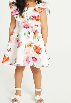 Robe blanche avec un imprimé floral vibrant en rose, orange et vert. Elle présente des manches volumineuses et une coupe évasée, associée à des sandales blanches.