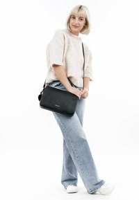 Bolso bandolera de cuero negro con un acabado texturizado, forma rectangular y cierre con cremallera, combinado con jeans de color azul claro y un top de punto beige.
