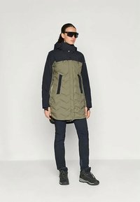 Πουκάμισο σε ελιά και ναυτικό μπλε, με σχέδιο ζιγκ-ζαγκ, τσέπες με φερμουάρ, ρυθμιζόμενη κουκούλα και σκούρα παντελόνια με μαύρες μπότες.