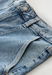 Detailní záběr modrých džín s páskem, poutek na opasek, kovovým knoflíkem s nápisem "NEXT DENIM" a podrobným prošíváním.