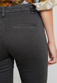 Närbild av gråa tajta byxor med en bakfickor med dragkedja och en liten svart Replay-logotyp, kombinerat med en uppkavlad ärm av en rutmönstrad skjorta.