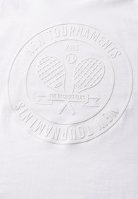 Bílý bavlněný tričko s vyvýšeným, tónovaným logem s tenisovými raketami, textem v kruhu "NOVÉ TURNÉ 2025" a bannerem "TENISOVÝ KLUB".