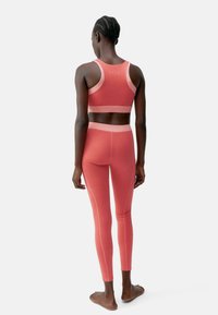 Sportos szett, amely egy testhez simuló, ujjtalan sportmelltartóból és magas derekú leggingsből áll, korall színben, ellentétes világos kiegészítőkkel és sima textúrával.