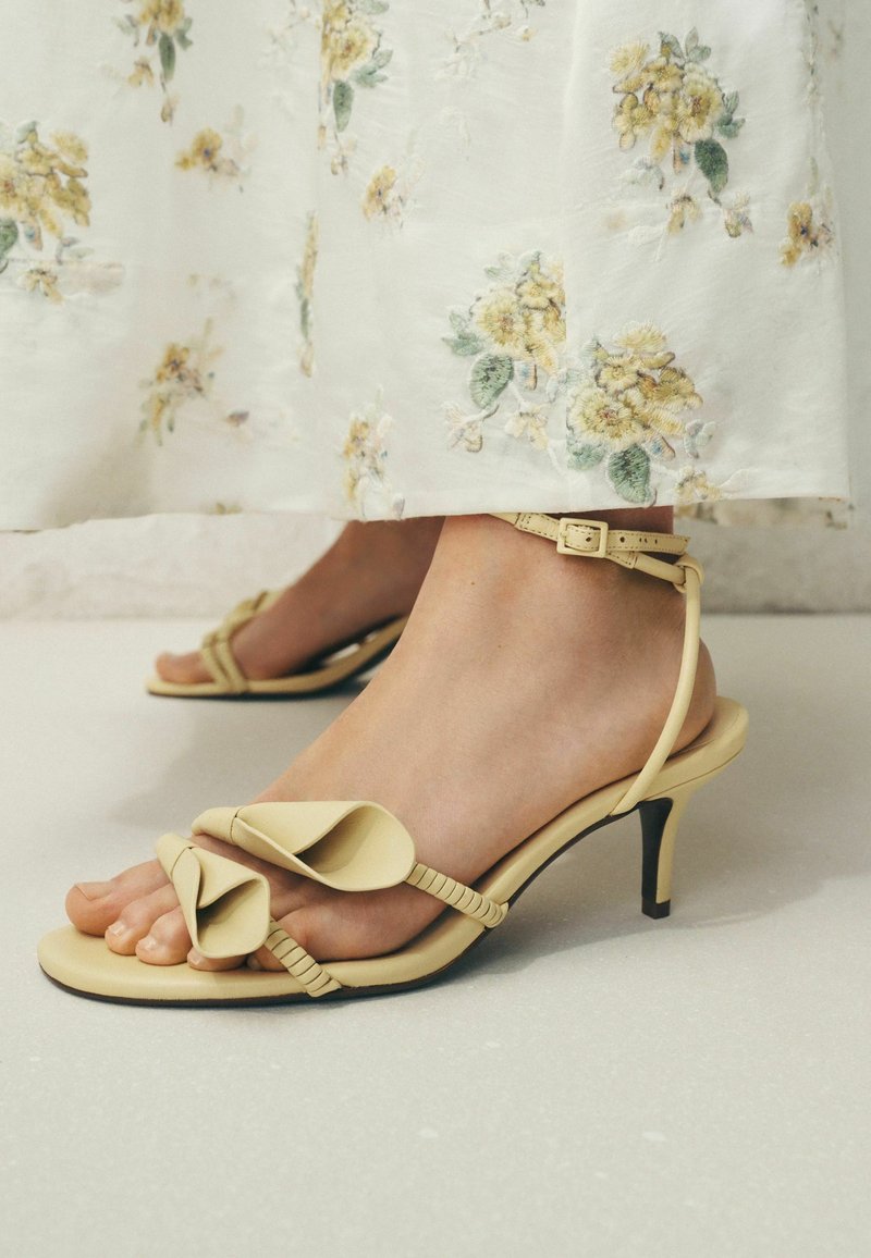 Sandales à talons beiges avec détail de bride torsadée, portées avec une robe blanche ornée d'une broderie florale jaune.